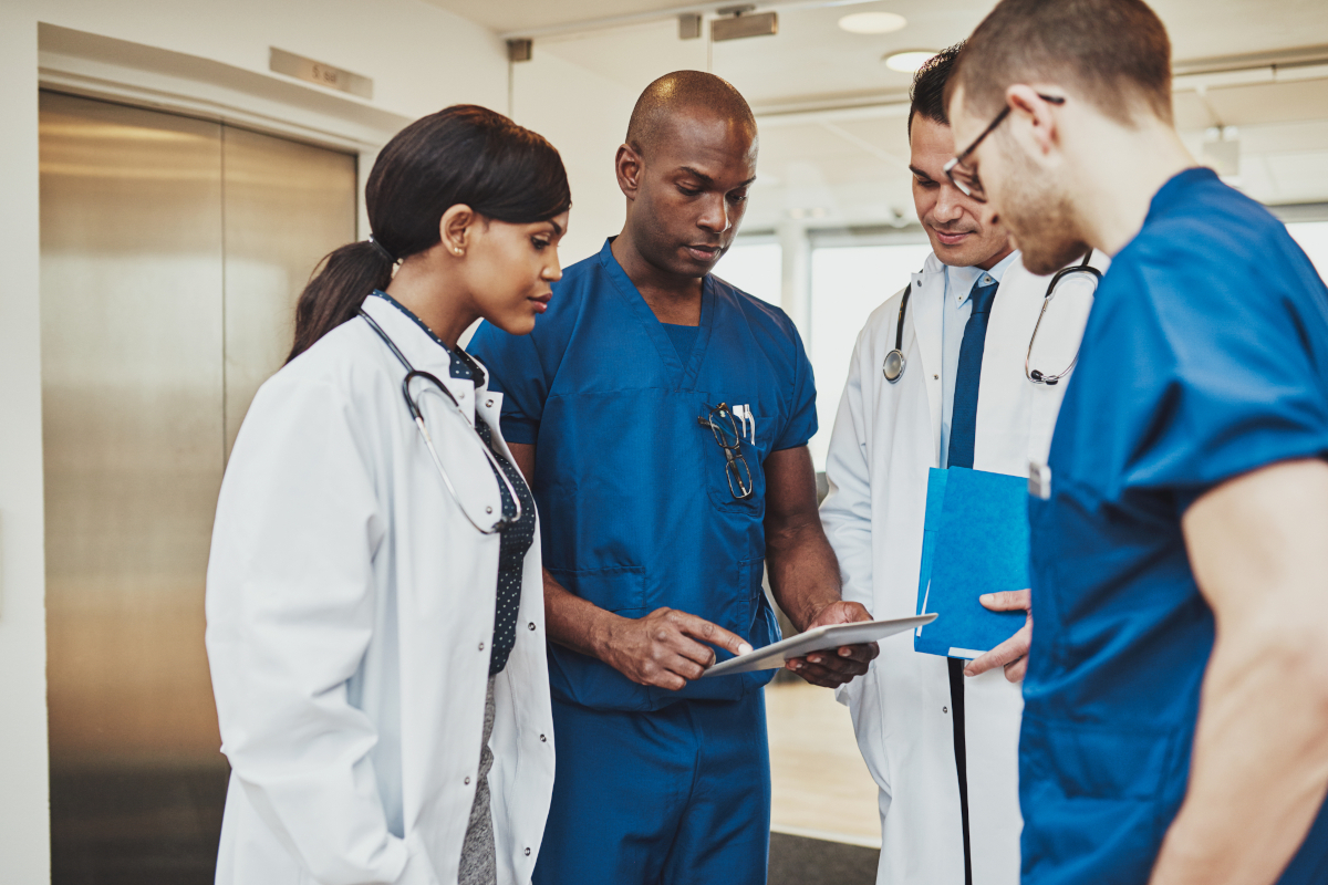 Four clinicians in scrubs and white coats reviewing a tablet together in a hospital hallway.