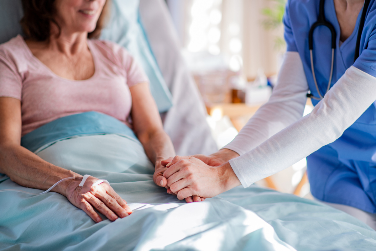 A clinician in blue scrubs holding the hand of a smiling patient resting in a hospital bed.